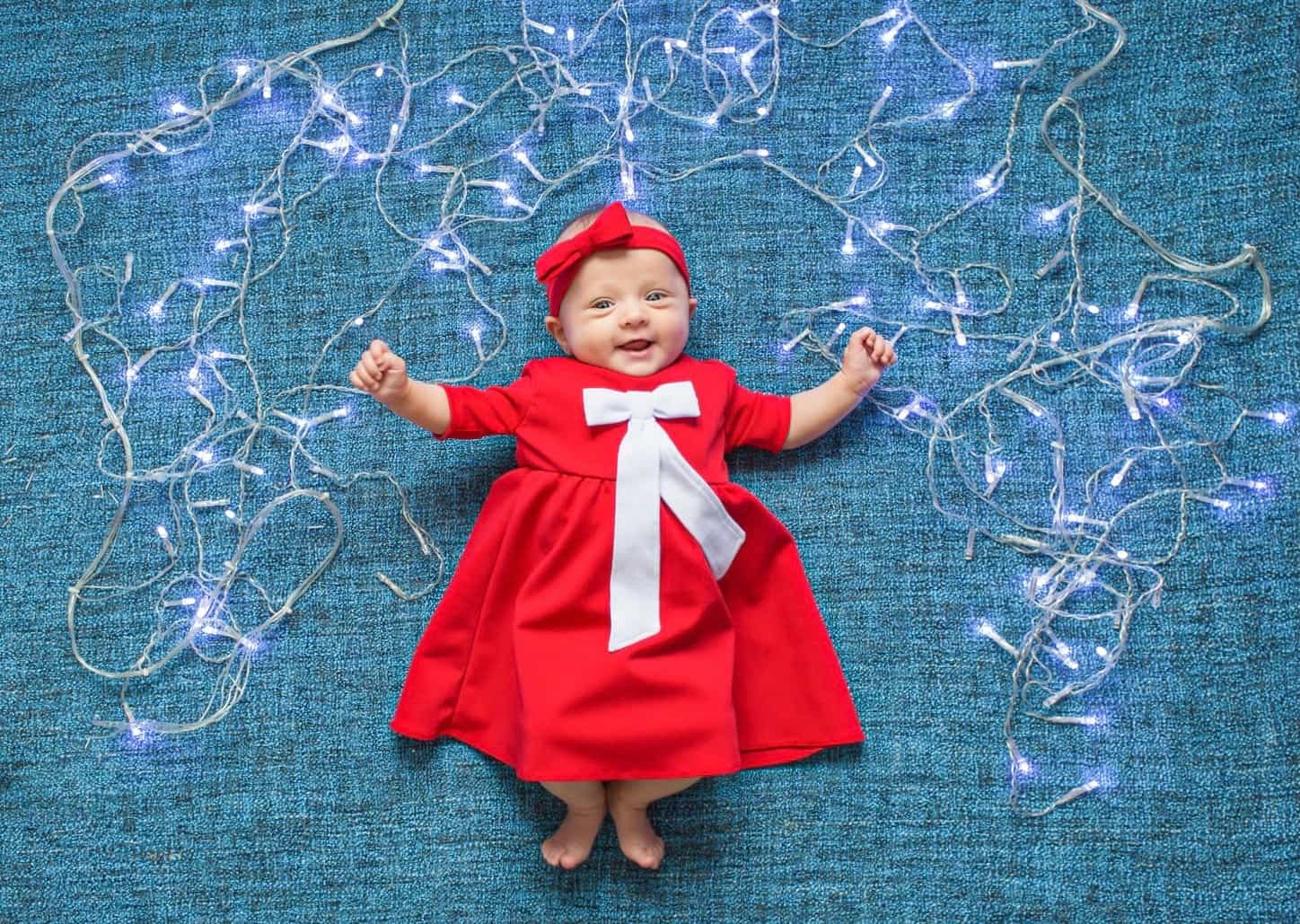 Infant in red dress lies and smiles on a blue background with Christmas lights