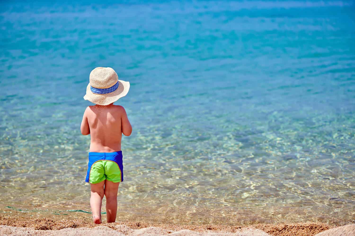 little boy standing in ocean water wearing hat